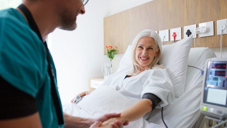 Male Nurse Taking Mature Female Patients Blood Pressure In Hospital Bed With Automated Machine