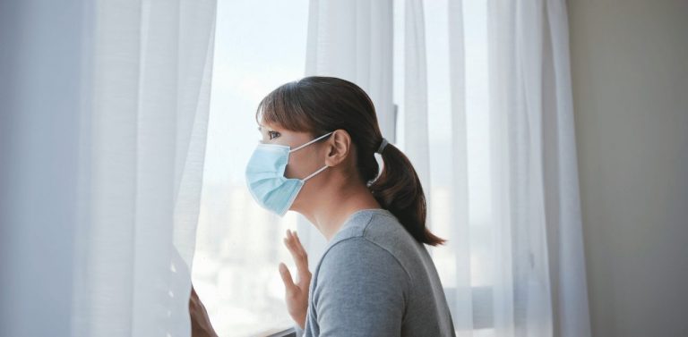 Woman in quarantine looking out window with mask on