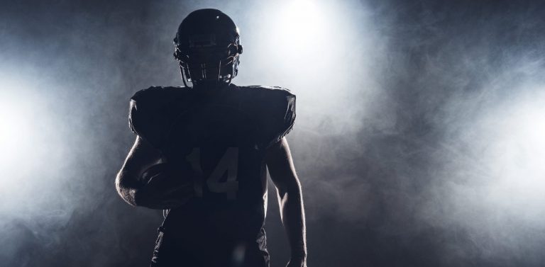 dark silhouette of equipped american football player with ball against white smoke